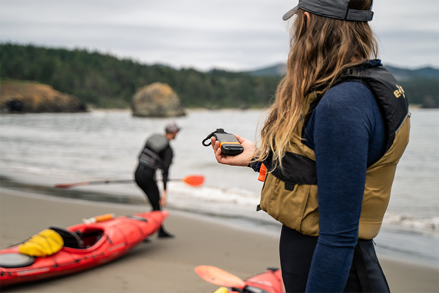 Two kayakers - one stands in the foreground holding a SPOT Satellite Messenger and the other is in the background pulling his kayak near the shore. 