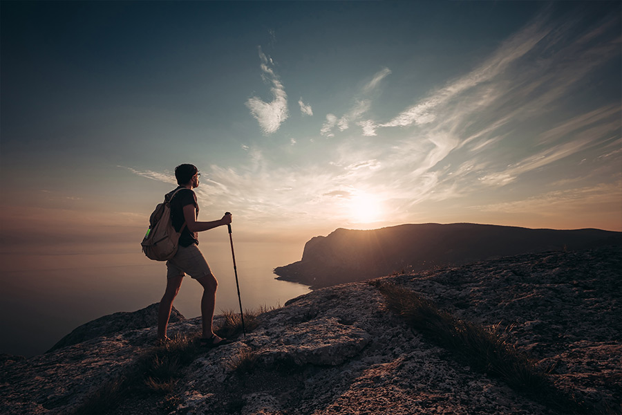 A lone male hiker holds a trekking pole as he reaches the summit of a coastal mountain at sunrise. He's wearing a backback with a ZOLEO messenger device attached to the top.