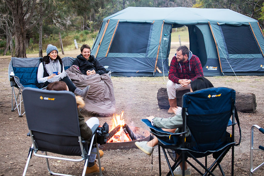 A group of campers gathered around a campfire in various OZtrail chairs, talking and laughing.