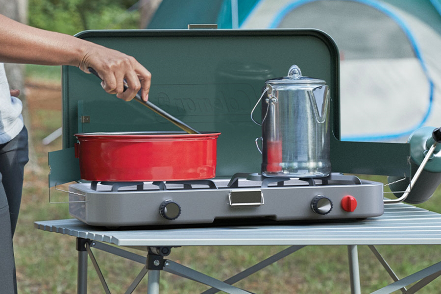 A hand stirring a red pot on a camp stove at a campsite. 