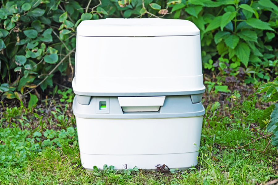 A portable toilet sitting on a green lawn in front of a green hedge.