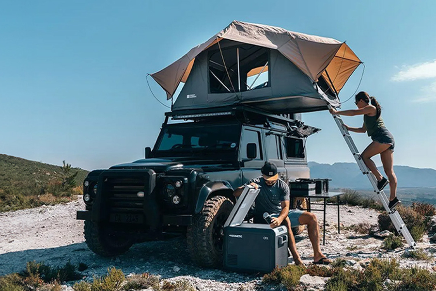 Two campers on a beach, one climbing the ladder up to the roof top tent on their 4WD, the other reaching into an esky.