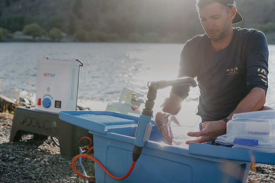 A man by a creek washing containers in a portable sink, with a Joolca HOTTAP to his right. 