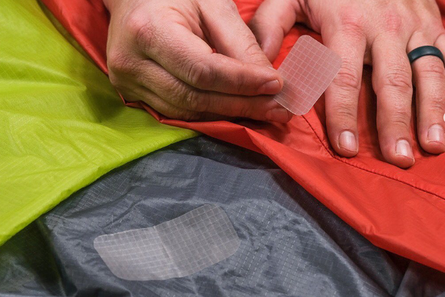 A close-up of a hand applying a repair patch to a green and orange tent. 