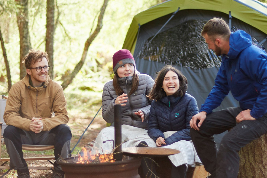 A group of campers talking and laughing around a campfire, rugged up in puffer jackets and rain coats. 