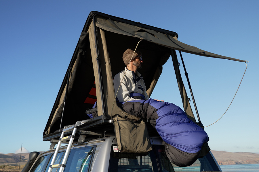 A man sitting up in his roof top tent, wearing a beanie and sunglasses, and wrapped in his sleeping bag. 