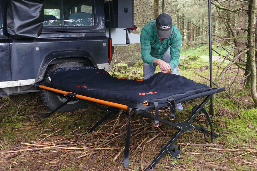 A man setting up a camp stretcher and sleeping bag at a campsite. 